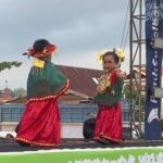 Two girls in bright traditional costumes with yellow flowers in their hair perform a cultural dance on an outdoor stage.