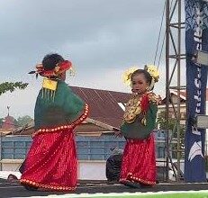 Two girls in bright traditional costumes with yellow flowers in their hair perform a cultural dance on an outdoor stage.