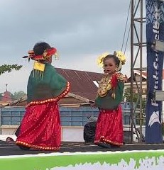 Two girls in bright traditional costumes with yellow flowers in their hair perform a cultural dance on an outdoor stage.