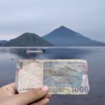 Hand holding a 1,000 Rupiah banknote in front of a calm lake with mountains and a boat in the distance (Indonesia)