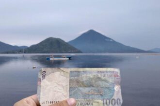 Hand holding a 1,000 Rupiah banknote in front of a calm lake with mountains and a boat in the distance (Indonesia)