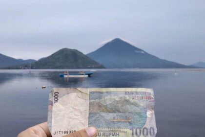 Hand holding a 1,000 Rupiah banknote in front of a calm lake with mountains and a boat in the distance (Indonesia)