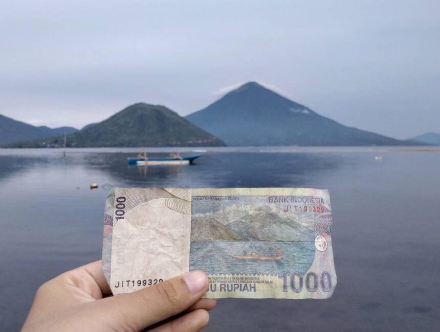 Hand holding a 1,000 Rupiah banknote in front of a calm lake with mountains and a boat in the distance (Indonesia)
