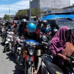 Line of motorcyclists on a city street, wearing helmets, with a woman in a purple hijab riding a scooter at the foreground.