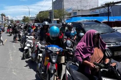 Line of motorcyclists on a city street, wearing helmets, with a woman in a purple hijab riding a scooter at the foreground.