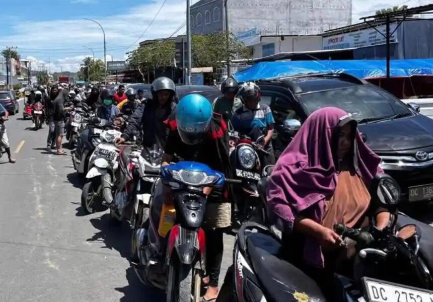 Line of motorcyclists on a city street, wearing helmets, with a woman in a purple hijab riding a scooter at the foreground.