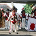 Historical reenactment: soldiers in white uniforms with plumed hats performing on a stage, carrying a white flag with a red star.