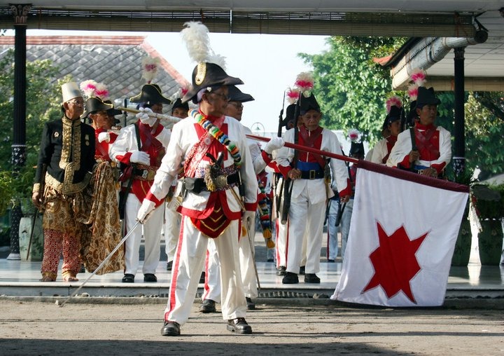 Historical reenactment: soldiers in white uniforms with plumed hats performing on a stage, carrying a white flag with a red star.