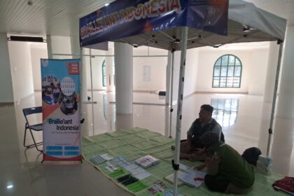 Informational booth for Braille Indonesia set up under a canopy in a bright hall, with banners, a folding chair, and brochures on a green mat for visitors with visual impairments.