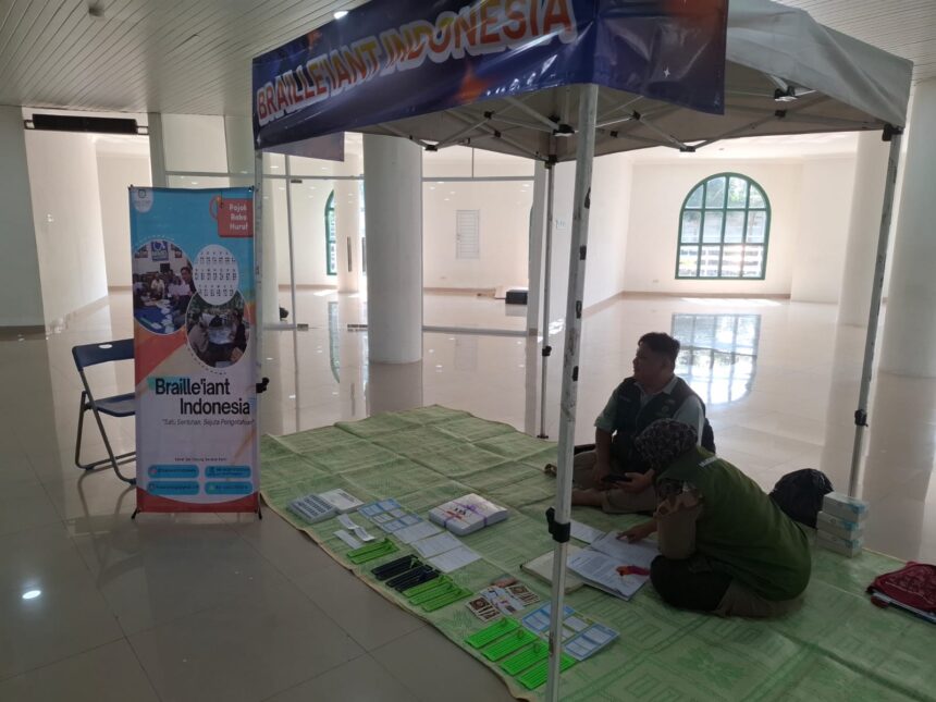 Informational booth for Braille Indonesia set up under a canopy in a bright hall, with banners, a folding chair, and brochures on a green mat for visitors with visual impairments.