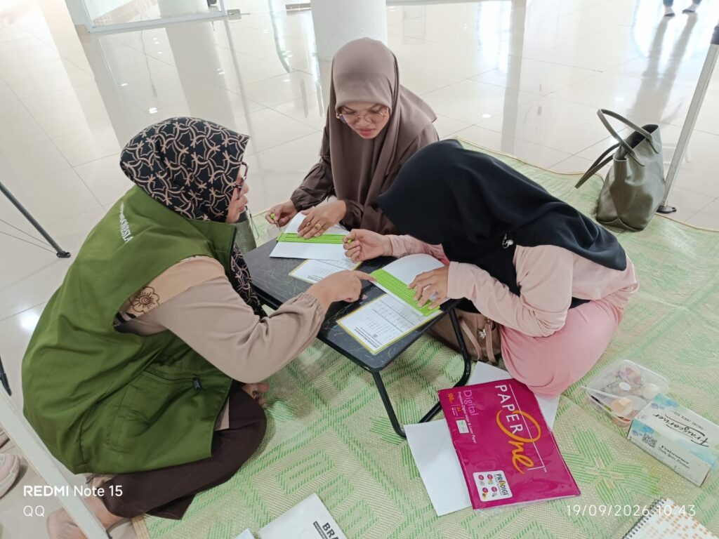 Three women in hijabs sit on a green mat around a small table, using bright green rulers to fill out documents in a bright, open space.