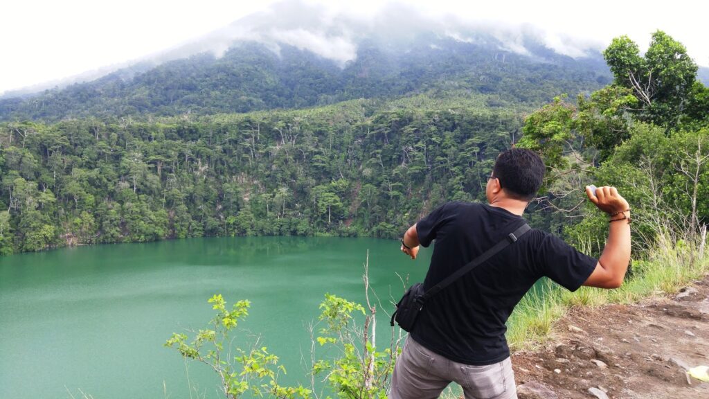 Man in a black shirt with a crossbody bag stands at a cliff, facing a green lake and dense forested mountains under cloudy skies.",