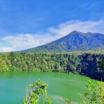 Vibrant green crater lake surrounded by dense forest and a blue sky with a distant volcanic peak.