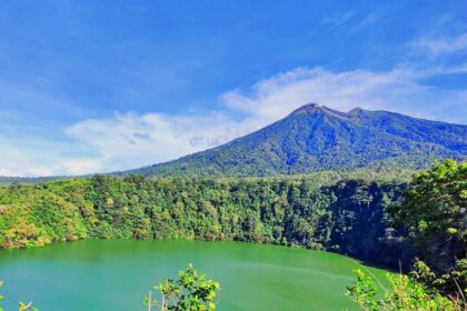 Vibrant green crater lake surrounded by dense forest and a blue sky with a distant volcanic peak.