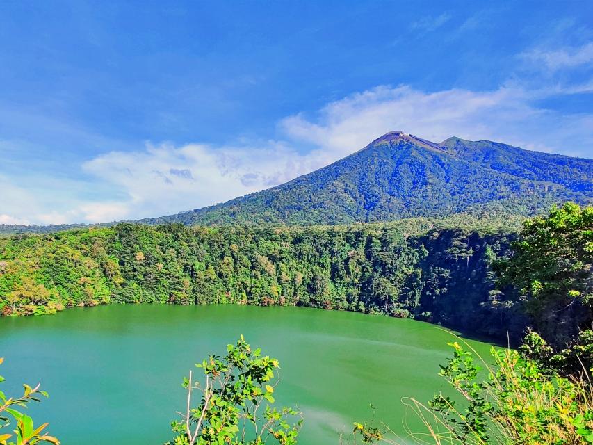Vibrant green crater lake surrounded by dense forest and a blue sky with a distant volcanic peak.
