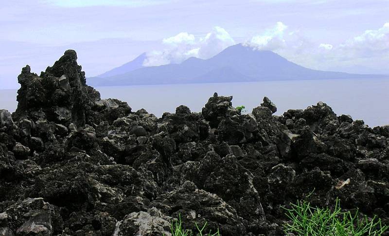 WhatsApp Image 2026-09-21 at 131952 - Mabur.co Rugged black lava rocks in the foreground, calm sea beyond, and a distant blue volcanic island under a partly cloudy sky.
