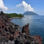 Rugged lava rocks along a calm blue sea with a distant island and small boats on the horizon.