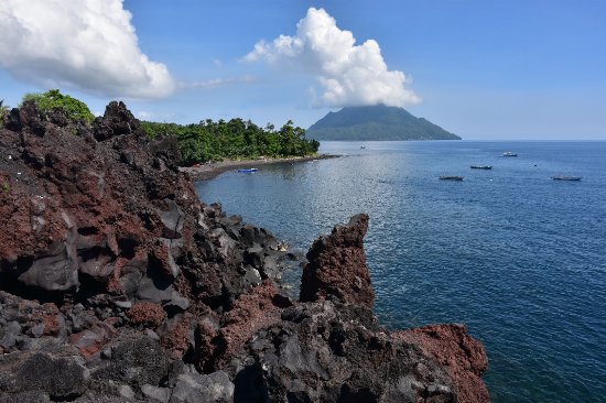 Rugged lava rocks along a calm blue sea with a distant island and small boats on the horizon.