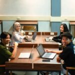 Four students sit around a wooden table in a classroom, working on laptops and smiling at the camera.