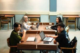 Four students sit around a wooden table in a classroom, working on laptops and smiling at the camera.