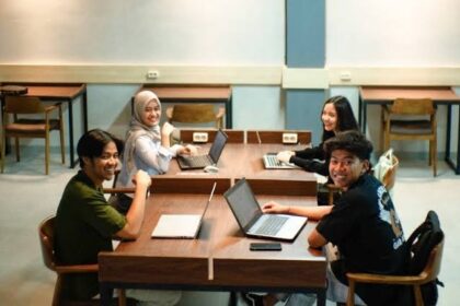Four students sit around a wooden table in a classroom, working on laptops and smiling at the camera.