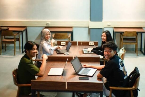 Four students sit around a wooden table in a classroom, working on laptops and smiling at the camera.