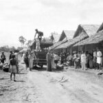 People gather along a dirt road in a thatched-hut village as a truck unloads goods and villagers watch.