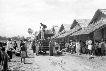 People gather along a dirt road in a thatched-hut village as a truck unloads goods and villagers watch.