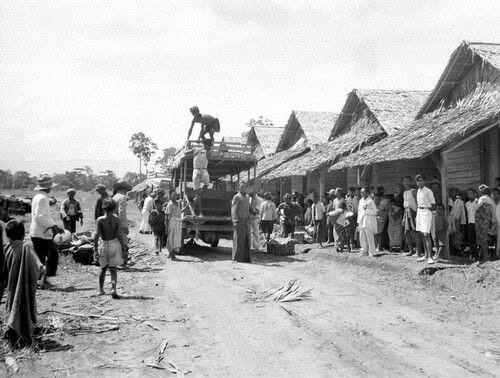 People gather along a dirt road in a thatched-hut village as a truck unloads goods and villagers watch.