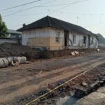 Old single-story building undergoing renovation, dirt road and construction materials in front, concrete pipes lined along the site.