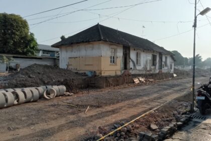 Old single-story building undergoing renovation, dirt road and construction materials in front, concrete pipes lined along the site.