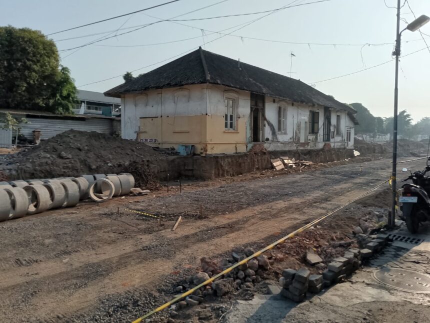 Old single-story building undergoing renovation, dirt road and construction materials in front, concrete pipes lined along the site.