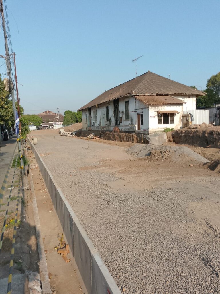 Construction site on a quiet street with a weathered white house, gravel and piles of dirt along the road under renovation.