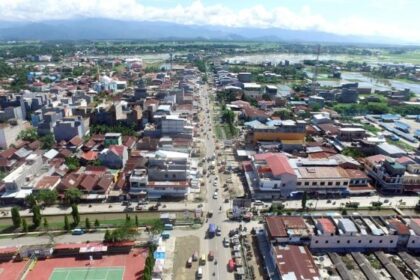 Aerial view of a bustling town with a central street lined by low-rise buildings, sports courts in the foreground, and distant hills on the horizon.