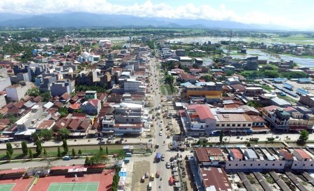 Aerial view of a bustling town with a central street lined by low-rise buildings, sports courts in the foreground, and distant hills on the horizon.