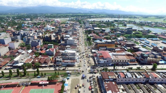 Aerial view of a bustling town with a central street lined by low-rise buildings, sports courts in the foreground, and distant hills on the horizon.