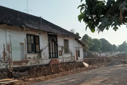 Old white house with cracked walls next to a dirt road under construction; ongoing foundation work and scattered wooden planks in the foreground
