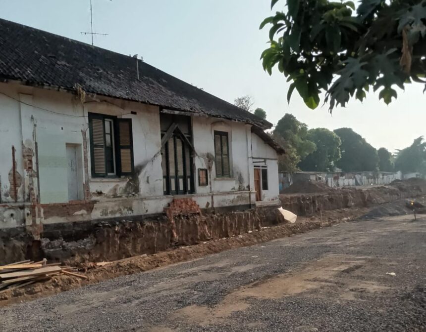 Old white house with cracked walls next to a dirt road under construction; ongoing foundation work and scattered wooden planks in the foreground