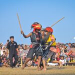 Two dancers with wooden staffs collide in a field while a large crowd watches from behind a rope barrier under a clear blue sky.