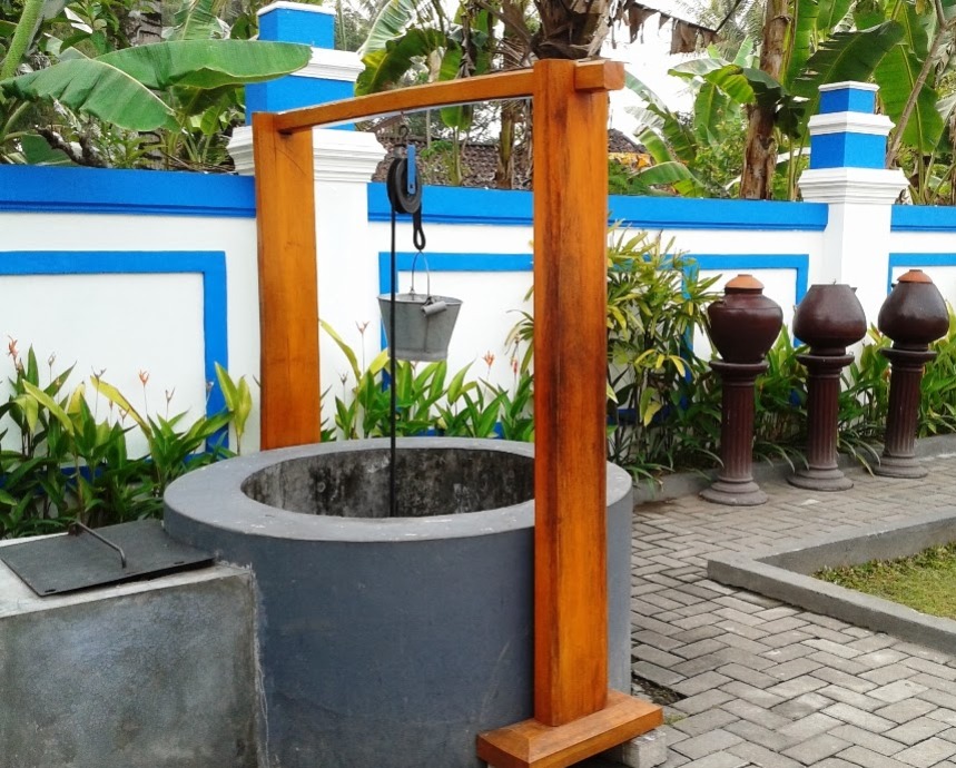 Wooden-framed hand-operated well with a hanging bucket in a landscaped courtyard, beside a blue-and-white wall and tropical plants.