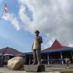 Bronze statue of a man in a suit standing in front of a blue building with a red-tiled roof and flagpole in a plaza.