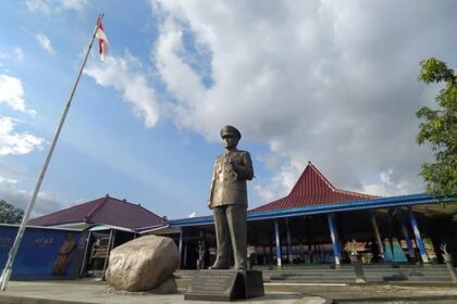 Bronze statue of a man in a suit standing in front of a blue building with a red-tiled roof and flagpole in a plaza.