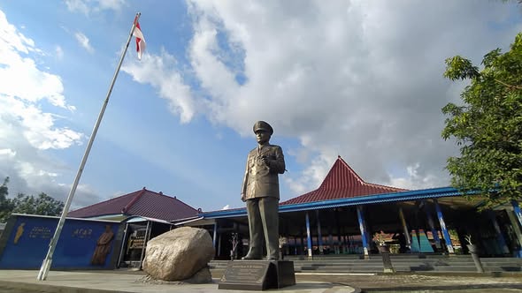 Bronze statue of a man in a suit standing in front of a blue building with a red-tiled roof and flagpole in a plaza.