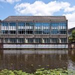Three-story concrete building with large window panels beside a canal; graffiti runs along the waterline wall, and lily pads float on the canal in the foreground.