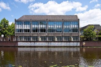 Three-story concrete building with large window panels beside a canal; graffiti runs along the waterline wall, and lily pads float on the canal in the foreground.