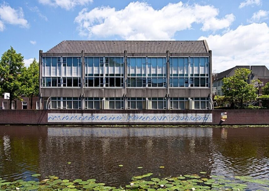 Three-story concrete building with large window panels beside a canal; graffiti runs along the waterline wall, and lily pads float on the canal in the foreground.