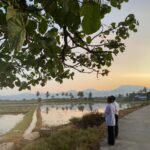 Two men in traditional clothing stand on a rural concrete road beside flooded rice fields at sunset, with palm trees and distant mountains in the background.