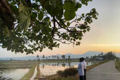 Two men in traditional clothing stand on a rural concrete road beside flooded rice fields at sunset, with palm trees and distant mountains in the background.