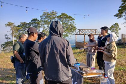 Group of people gathered around a table outdoors on a hillside, presenting or discussing items as a wooden cart backdrop sits nearby.