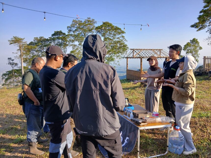 Group of people gathered around a table outdoors on a hillside, presenting or discussing items as a wooden cart backdrop sits nearby.